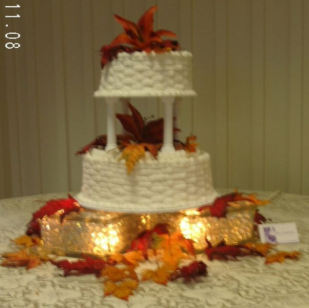 Two-tiered wedding cake with white textured frosting, autumn leaves, and orange flowers. Lit-up base.