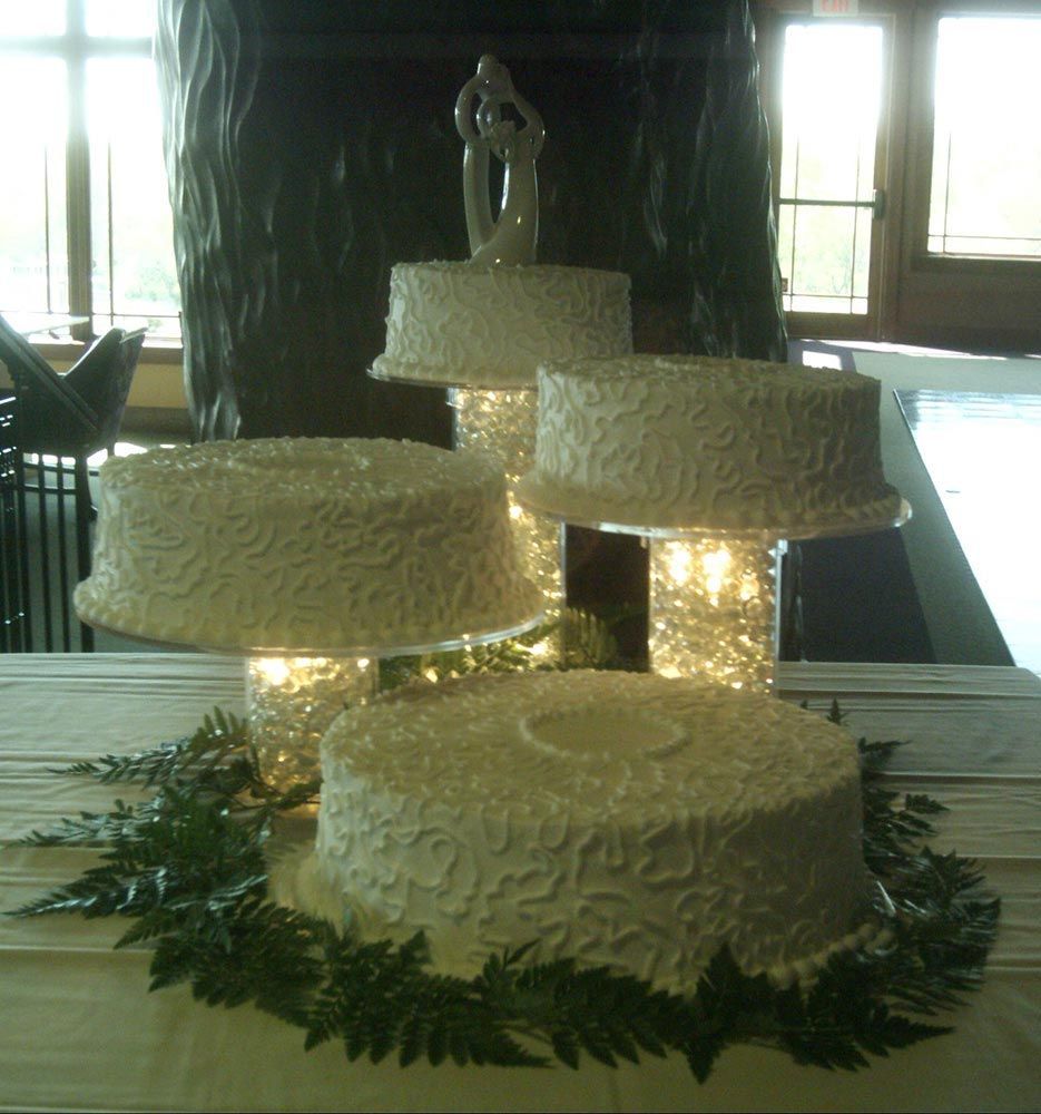 Four-tiered wedding cake with white textured frosting, topped with a bride and groom figurine. Lit pedestals.