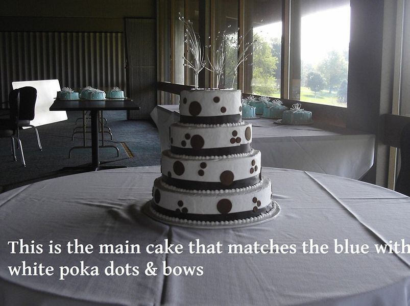 Four-tiered white cake with brown polka dots and bows; matching blue cupcakes on tables in background.