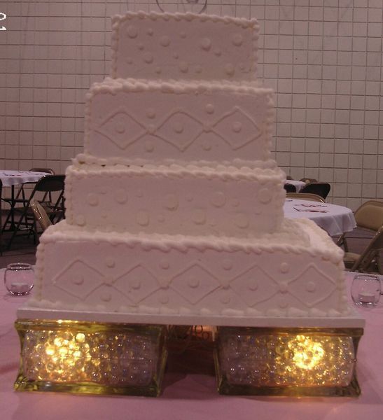 Four-tiered white wedding cake with diamond and heart details, resting on a glowing, ornate base.