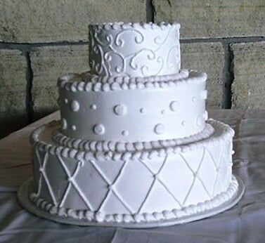 Three-tiered white wedding cake decorated with swirls, dots, and diamond patterns.