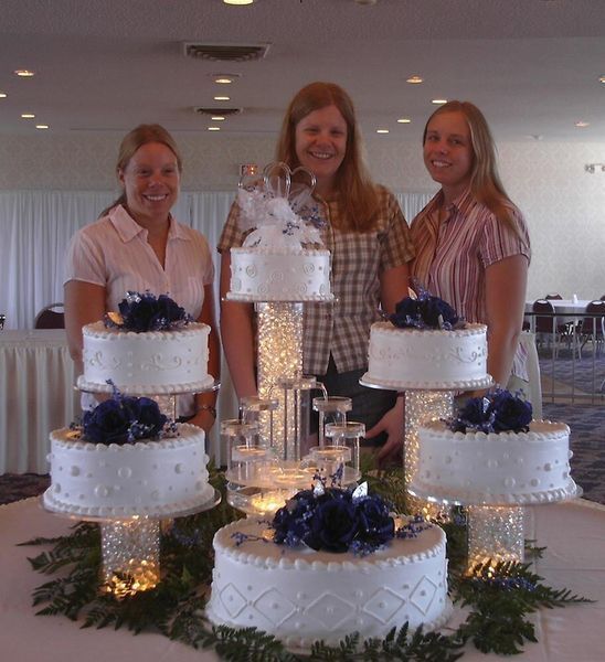 Three women stand behind a tiered wedding cake display with blue flower accents.