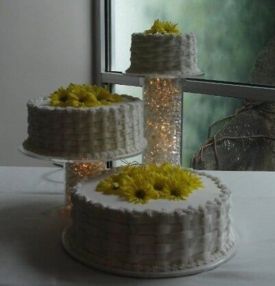 Three-tiered cake with white frosting and yellow flower decorations, on stands in front of a window.