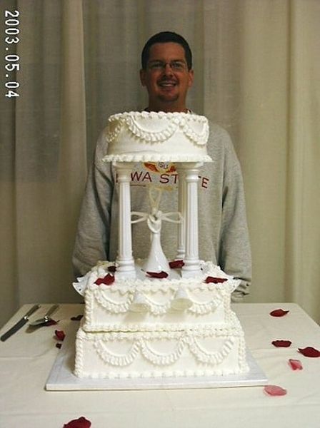 Man smiling behind a tiered white wedding cake with red rose petals, set on a white tablecloth.