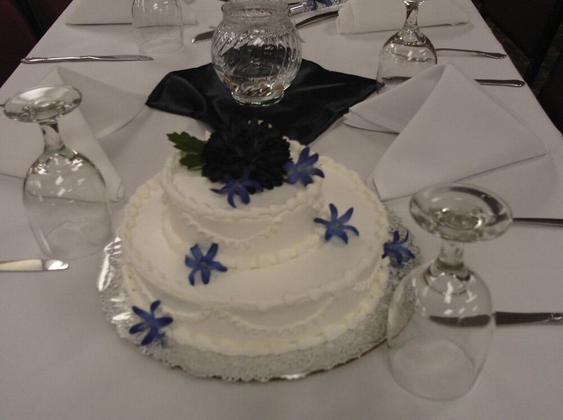 Cake on a table, decorated with blue flowers, surrounded by glassware and silverware, likely at a formal event.