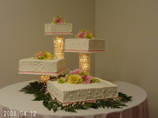 Four-tiered wedding cake with square layers, decorated with flowers, lit from within, on a round table.