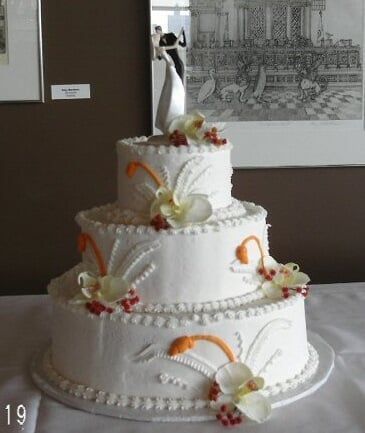 Three-tiered white wedding cake decorated with orange and white floral accents, topped with bride and groom figurines.
