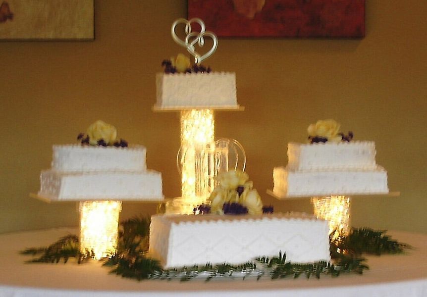 Wedding cake display with four tiers, white icing, adorned with flowers, on illuminated pedestals.