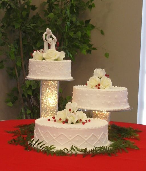 Wedding cakes on tiered stands, decorated with white roses and a bride and groom topper, on a red tablecloth.