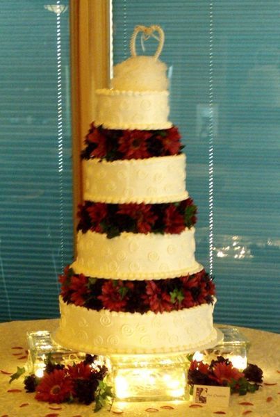 Five-tiered white wedding cake decorated with red flowers, topped with heart-shaped decoration, on a table.