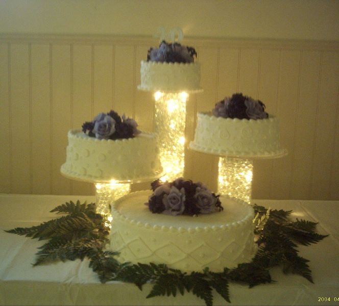 Four-tiered wedding cake with purple flowers, on lighted pedestals, on a white tablecloth with greenery.