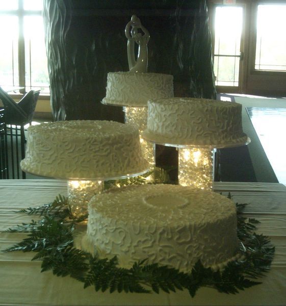 Four-tiered wedding cake with white frosting and a couple topper, surrounded by greenery and lights.