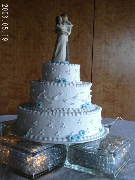 Three-tiered wedding cake with white frosting, blue flowers, and a couple figurine on top.