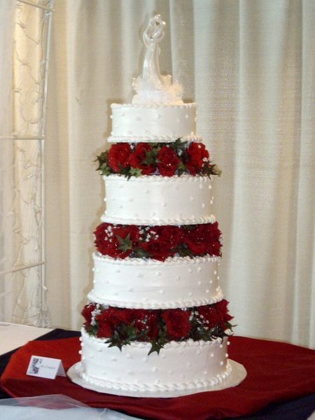 Wedding cake with five tiers, white frosting, red roses, and a bride and groom topper.