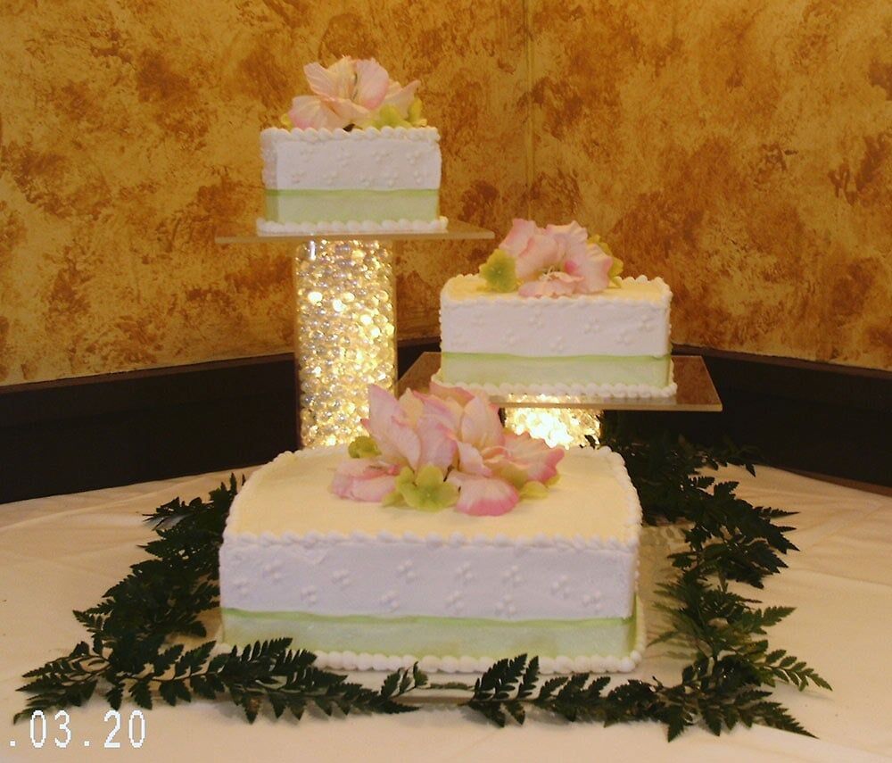 Three-tiered wedding cake display with pink and green floral accents on white frosting, with a warm-toned background.