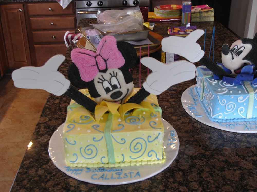 Minnie Mouse cake emerging from yellow gift box with pink bow. Another Mickey Mouse cake nearby.