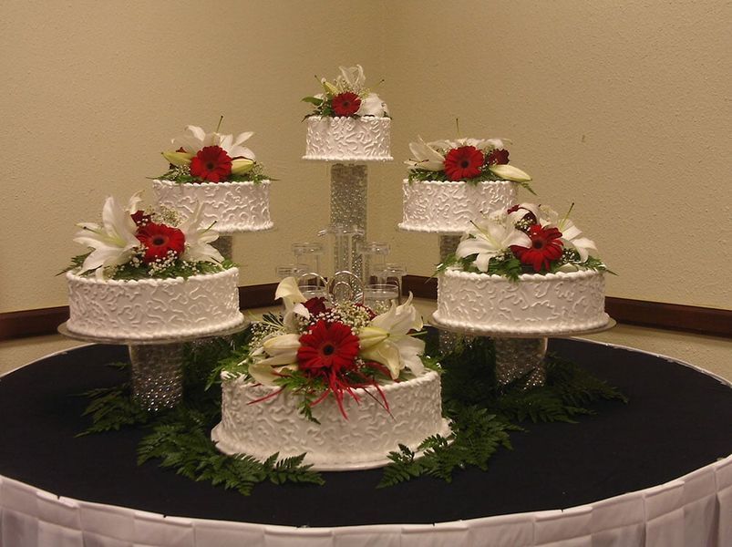 Tiered wedding cake display with white frosting, red flowers, and lilies, set on a dark tablecloth.