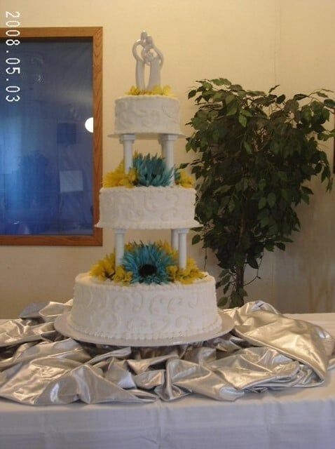 Four-tiered white wedding cake with blue and yellow flower accents, a silver topper, on a draped silver cloth.