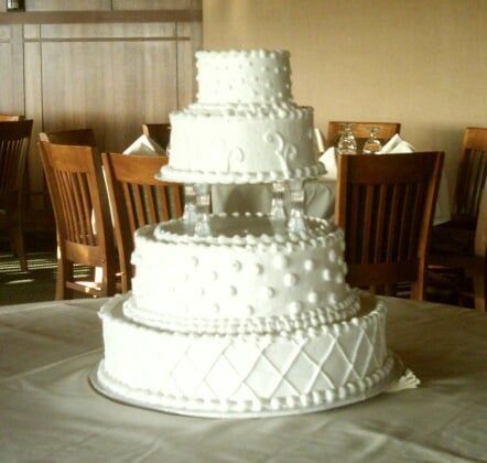 Four-tiered white wedding cake with intricate decorations on a table in a dining room.