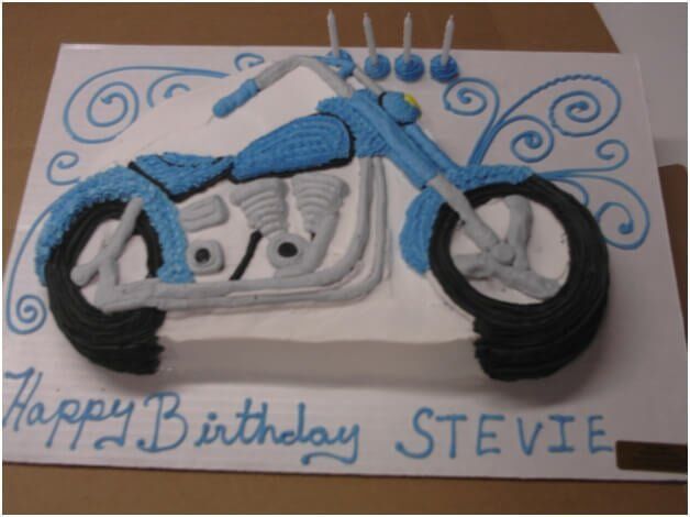 Birthday cake decorated as a blue motorcycle with gray engine, black tires, and blue cursive 