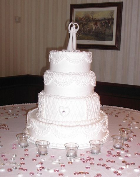 Four-tiered white wedding cake with couple figurine on top, surrounded by candles and pink confetti on a table.