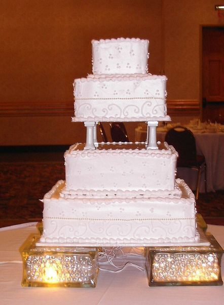 Four-tiered white wedding cake with ornate frosting and a lit base, set in a banquet hall.