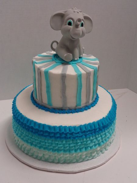 Two-tiered cake with a baby elephant topper. Bottom tier has blue ombre ruffles, top tier is striped.