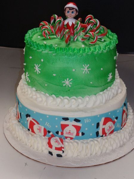 Two-tiered Christmas cake: green frosting, snowflakes, Santa ribbon, candy canes, and Elf on the Shelf figurine on top.