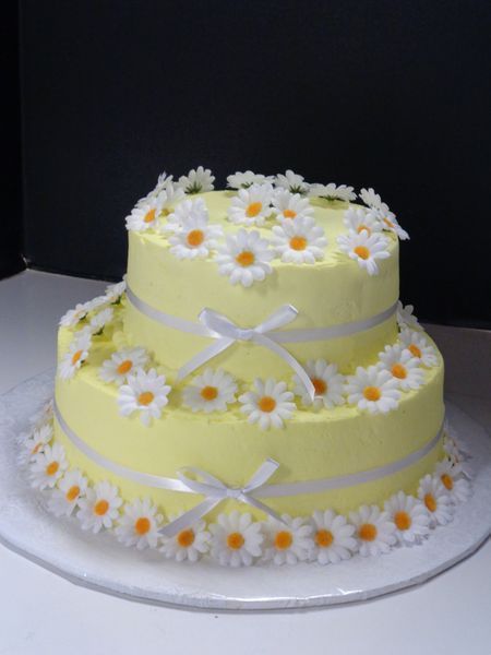 Two-tiered yellow cake decorated with white daisies and ribbon bows on a white plate.