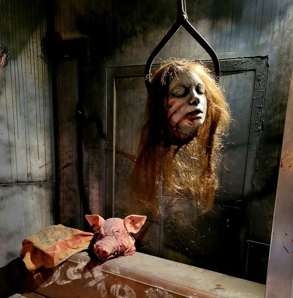 Hanging bloody mask in a grimy room, with a pig head on a table in the foreground.