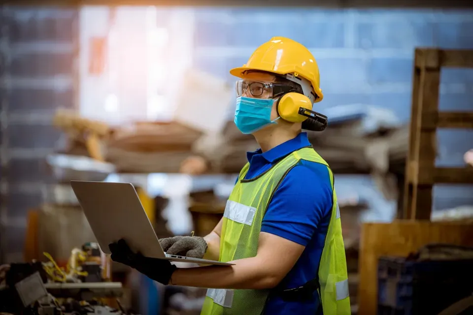 man in workplace setting, wearing mask and hardhat, using laptop computer