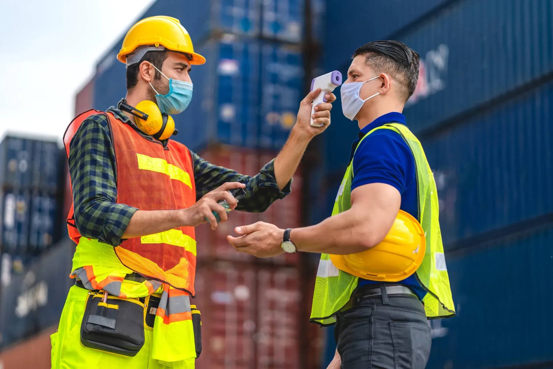 man taking temperature of other man in workplace setting, wearing masks and hardhats