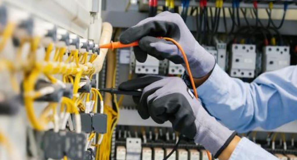 Technician in gloves testing an electrical panel with an orange probe and black meter leads.