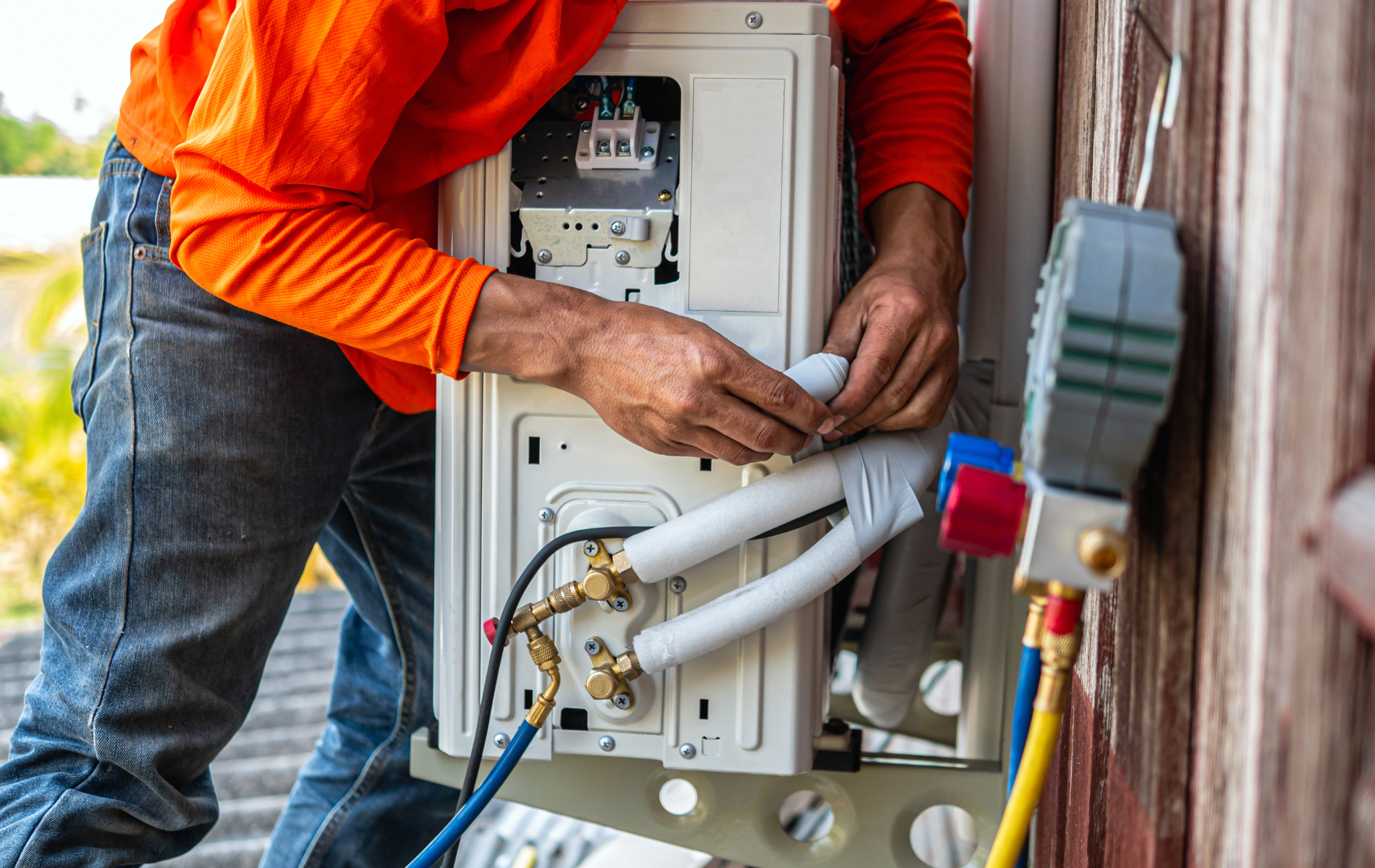 Technician in orange shirt inspecting outdoor HVAC unit with exposed wiring and pipes