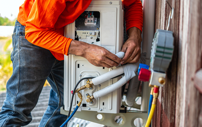 Technician in orange shirt inspecting outdoor HVAC unit with exposed wiring and pipes