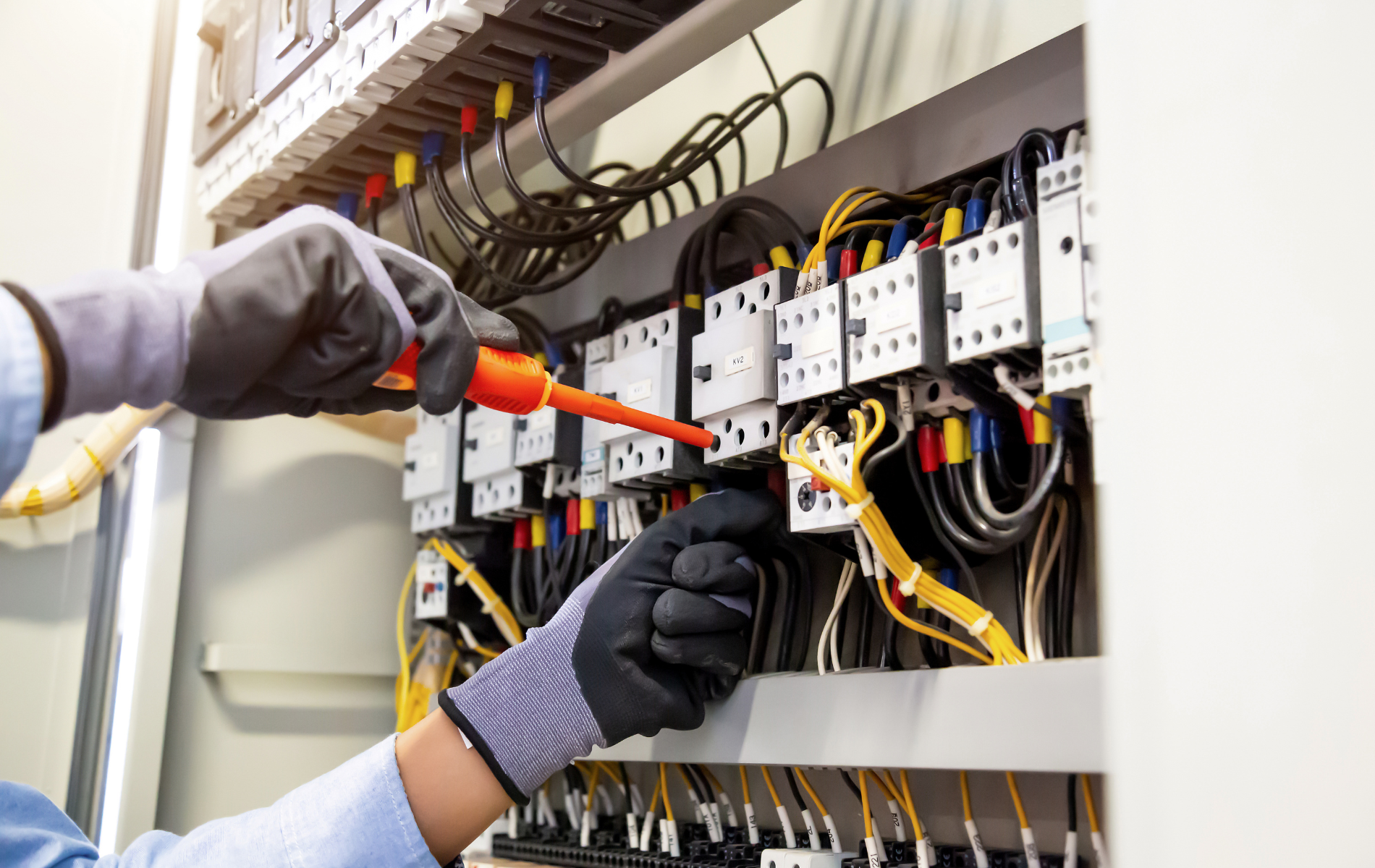 Technician wearing gloves inspecting wiring inside an electrical control panel with a screwdriver