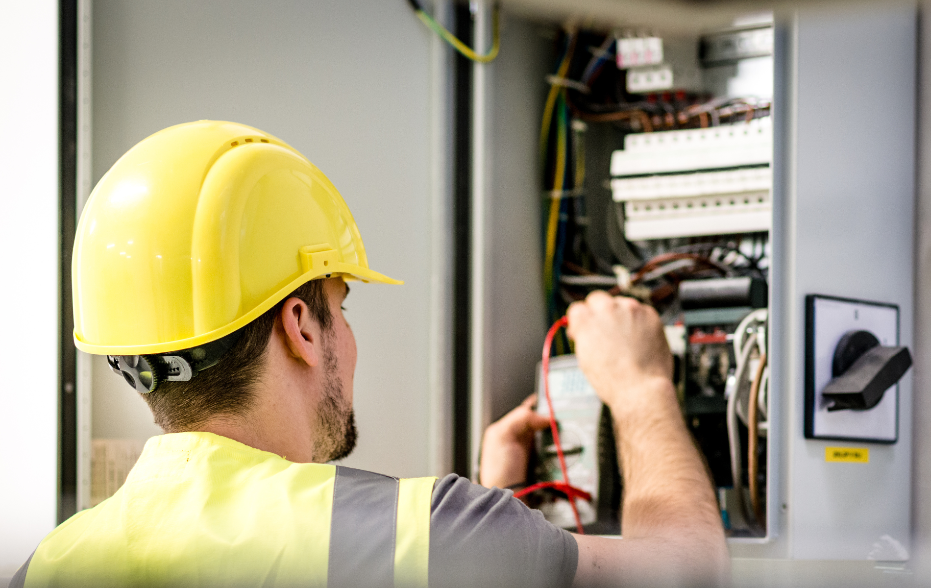 Technician in yellow hard hat inspecting electrical control panel with wires exposed