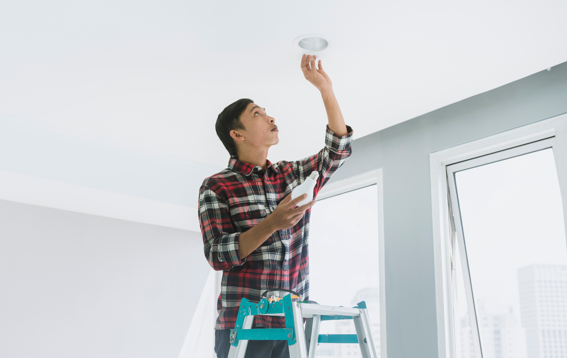Man on a ladder installing a ceiling light in a bright room with white walls and windows