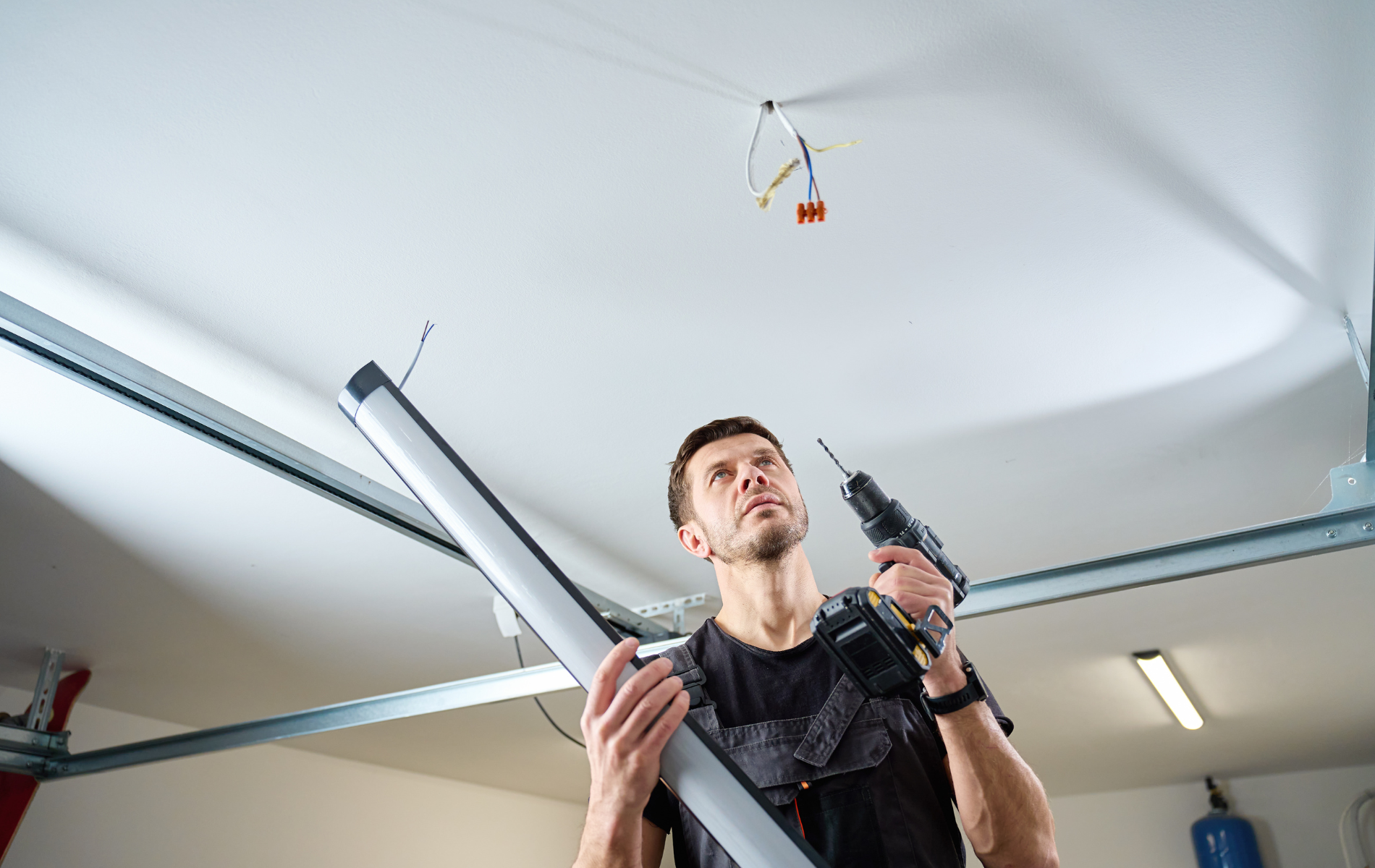 Man installing ceiling fixtures, holding a drill and long metal pipe in a garage-like room