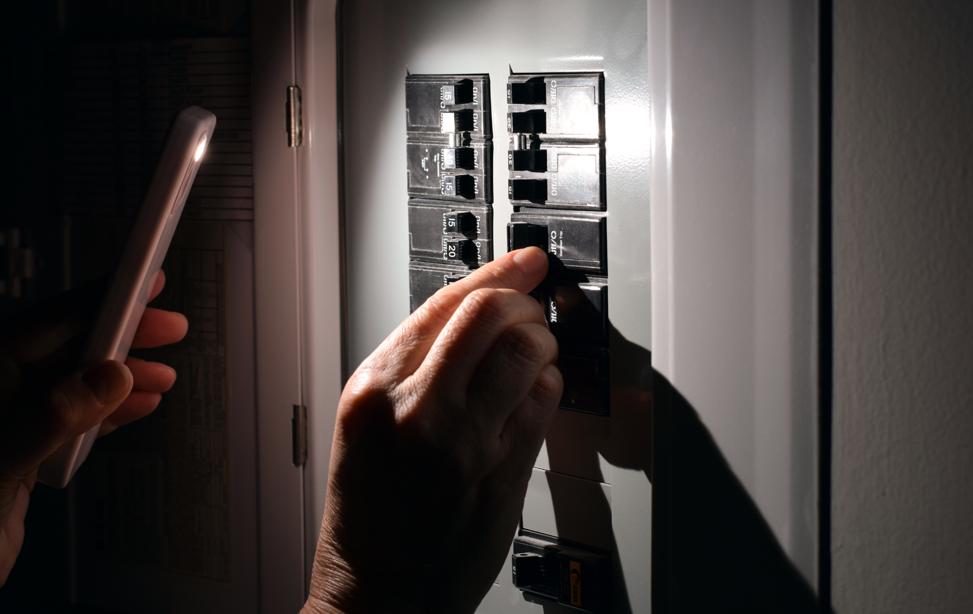 Hands inspecting an electrical breaker panel with a flashlight in a dim room