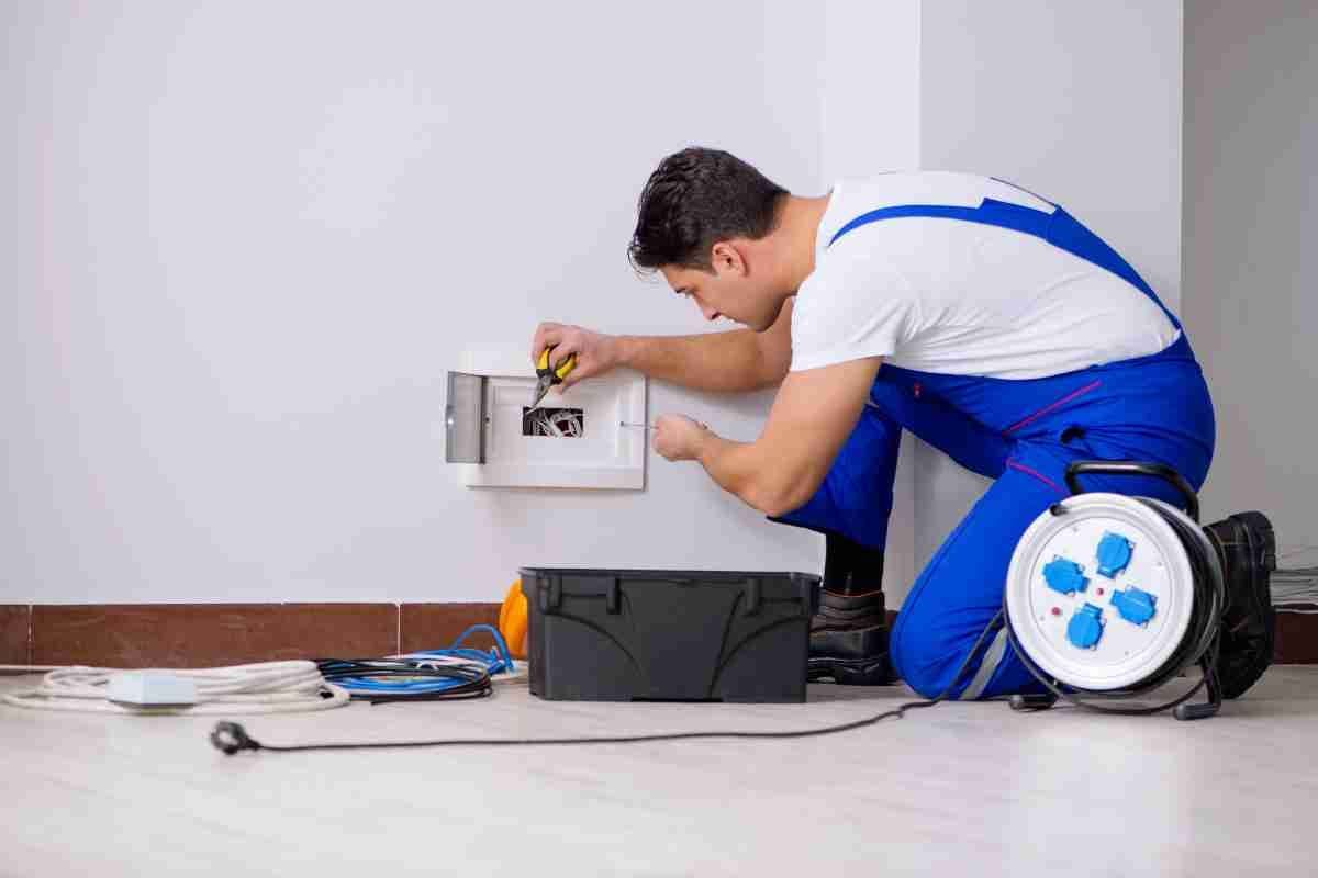Electrician kneeling by a wall outlet, working on wiring beside tools and a cable reel.