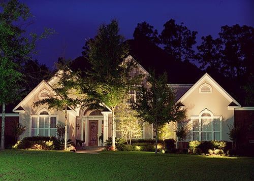 Well-lit suburban house at night with trees and a green lawn