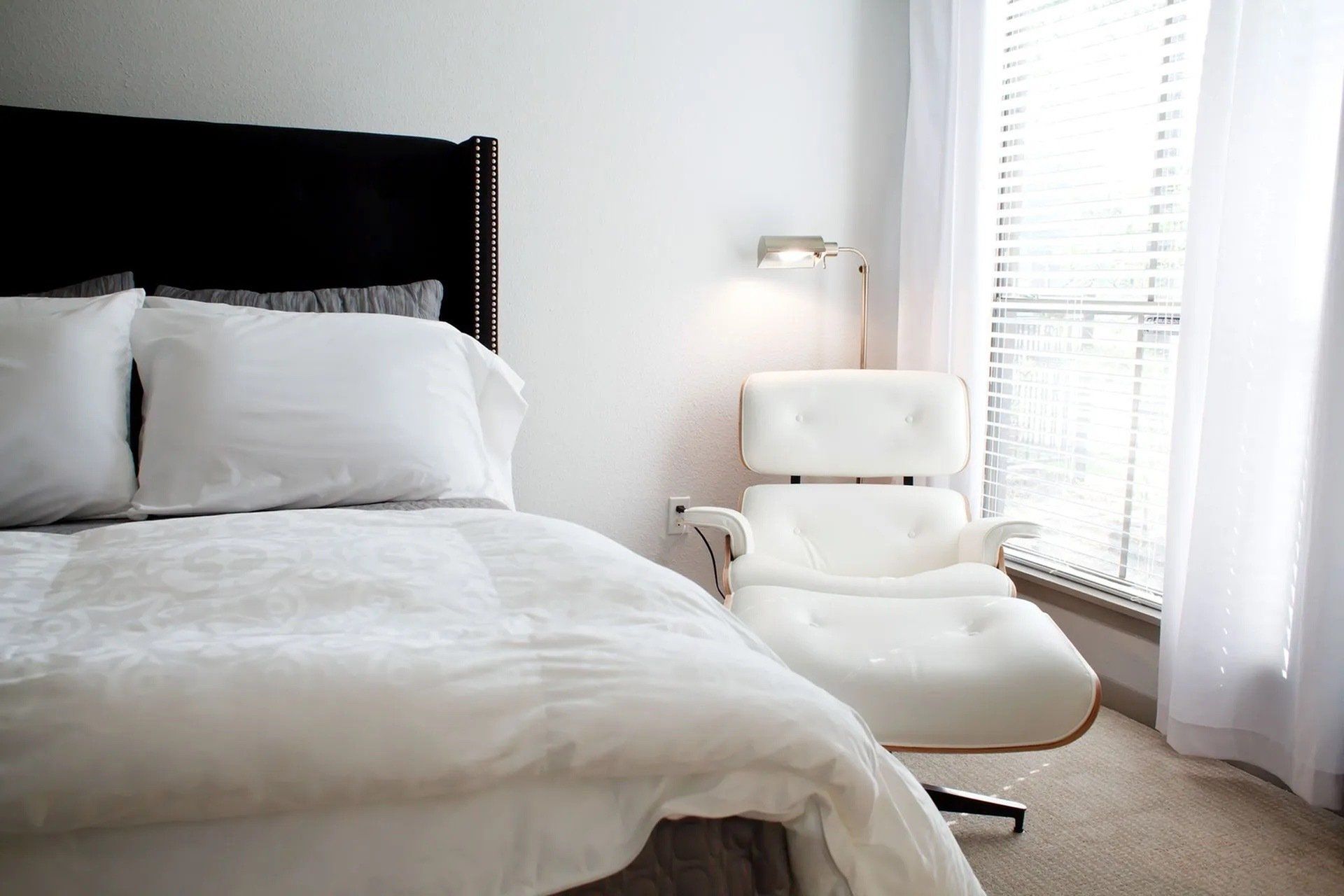 Bright bedroom with white bedding and a white lounge chair by a window.