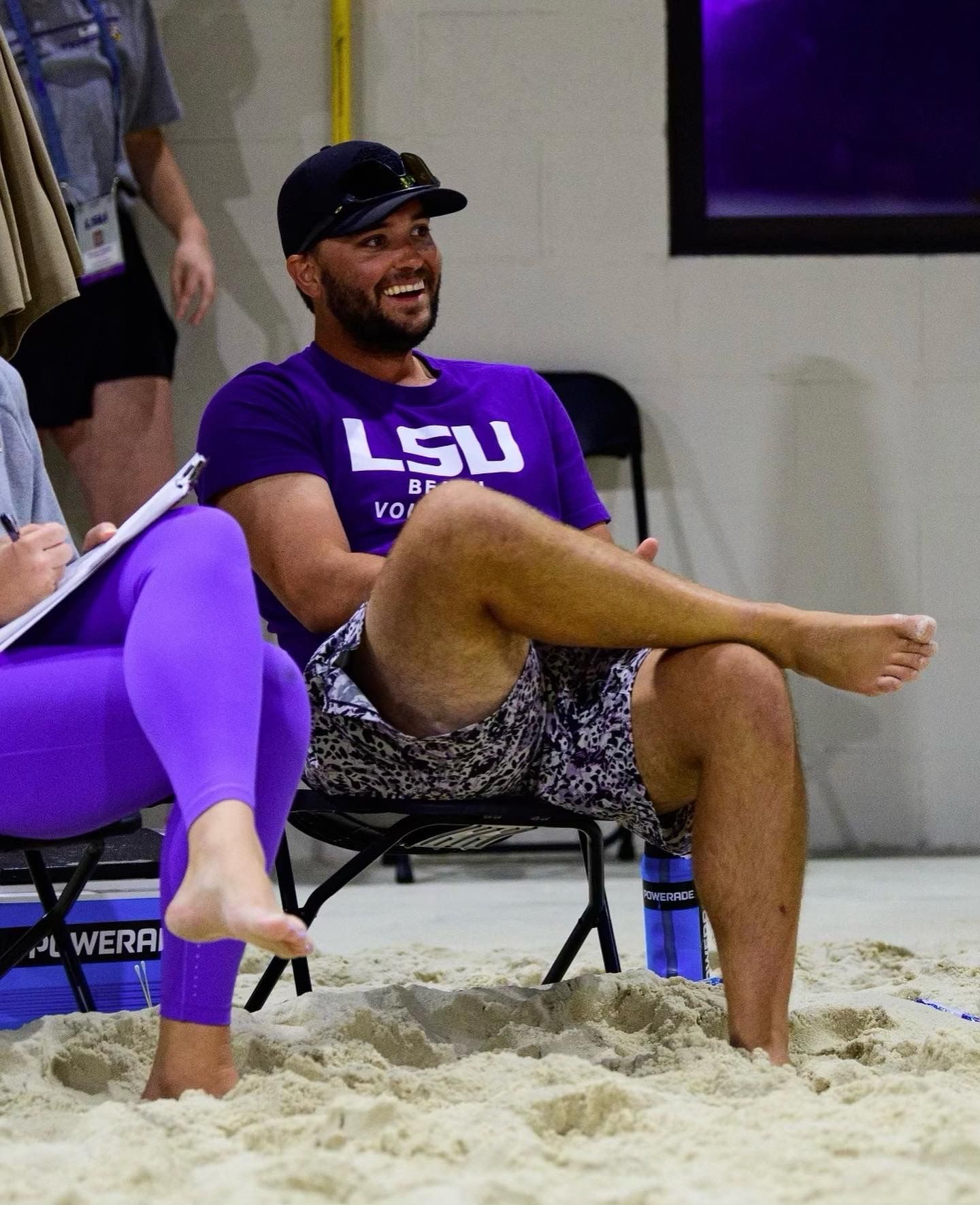 Man smiling on a sand court, wearing a purple LSU shirt and black cap, seated with legs crossed.