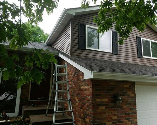Ladder leaning against a brick house under roof repair, with gray siding and green trees overhead