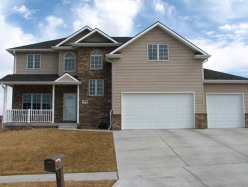 Suburban two-story house with beige siding, stone facade, white garage doors, and a front porch.