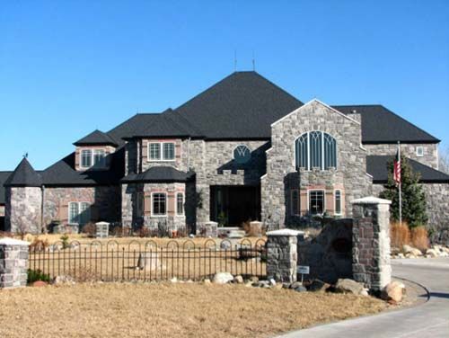 Large stone house with dark roof, arched front entrance, and a low stone wall under a clear blue sky