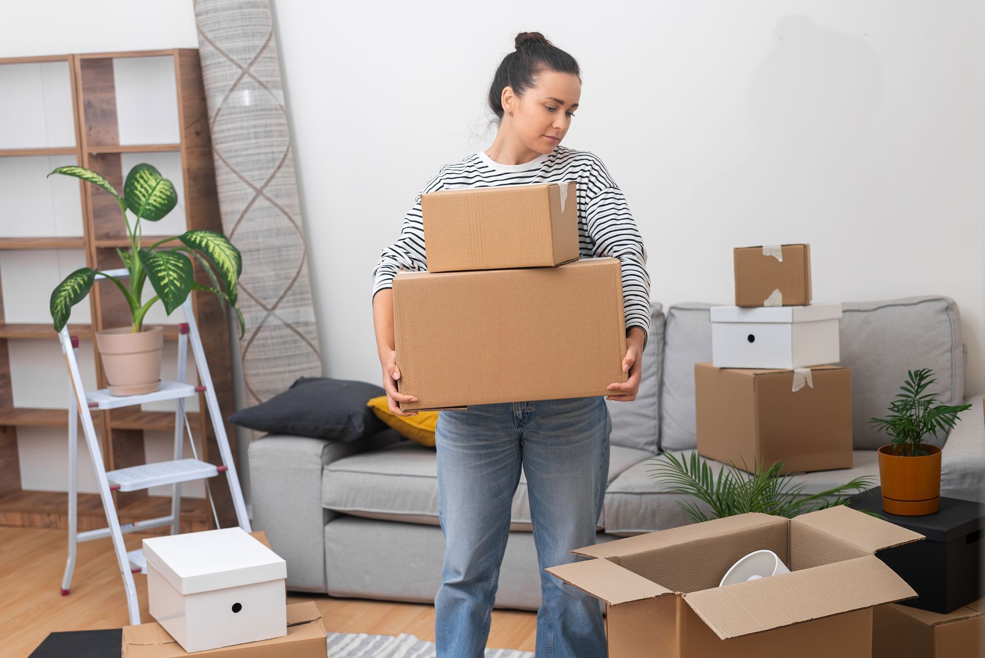 Woman holding two cardboard boxes while moving. Boxes and furniture in the background.