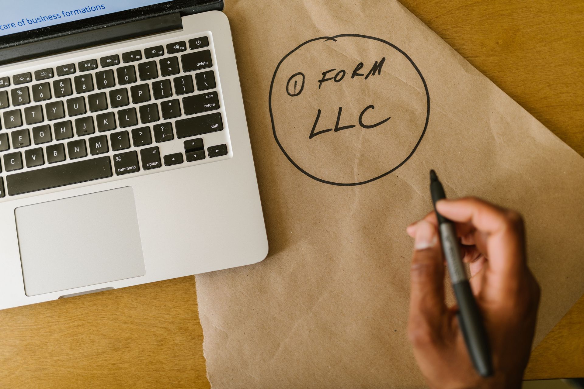 Hand writing “LLC” on brown paper beside a laptop