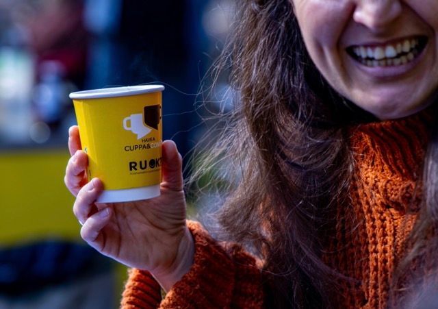 A smiling woman with long dark hair holding a yellow R U OK? coffee cup
